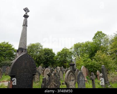 Cimitero storico a Manchester, Regno Unito, con lapidi erose, un alto monumento a croce e alberi verdi rigogliosi. Foto Stock