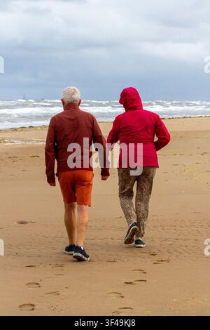 Vista posteriore di una coppia anziana in cappotti caldi che cammina lungo una spiaggia sabbiosa verso un mare agitato e ventoso in una giornata nuvolosa. Southport, Regno Unito. Foto Stock