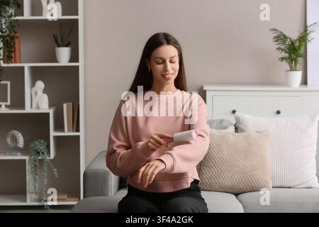 Giovane donna che usa un rullo anti-pelucchi sulla sua felpa rosa mentre è seduta su un divano grigio in un soggiorno moderno Foto Stock