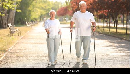 Coppia di anziani felici che fa Nordic Walking con i bastoni in un parco soleggiato, promuovendo uno stile di vita attivo e un invecchiamento sano, l'uomo e la donna anziani sorridono Foto Stock