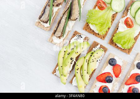 Set di sandwich stagionali biologici su fette di pane integrale marrone secco con verdure fresche, frutti maturi, conserve di pesce, formaggio fresco su tavolo di legno bianco, vista dall'alto, bordo. Foto Stock