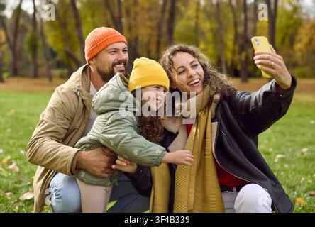 Genitori con figlia che scatta un selfie in un parco autunnale, famiglia felice all'aperto Foto Stock