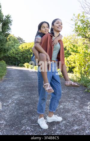 Madre afroamericana che porta sulla schiena la figlia adolescente camminando lungo un sentiero lastricato del parco, oltrepassando dei cespugli Foto Stock