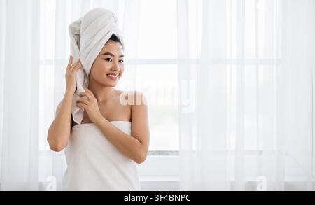 Donna asiatica avvolta in asciugamano in piedi accanto alla finestra Foto Stock