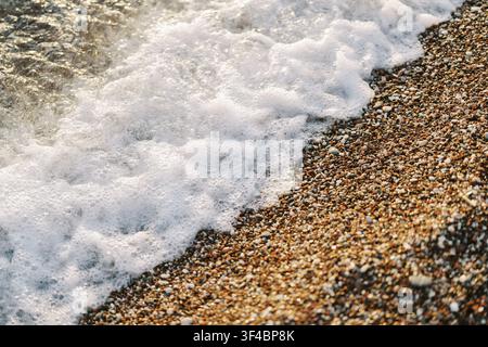 Spiaggia di ciottoli con onde schiumose che fanno brillare pietre multicolori bagnate dalla dolce marea lungo la costa tranquilla, texture in primo piano, luce soffusa Foto Stock