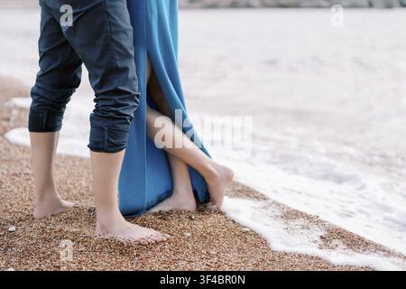 Una vista ravvicinata di una coppia in piedi a piedi nudi su una spiaggia di ciottoli, con onde gentili che lambiscono i loro piedi. La donna indossa un vestito blu con un Foto Stock