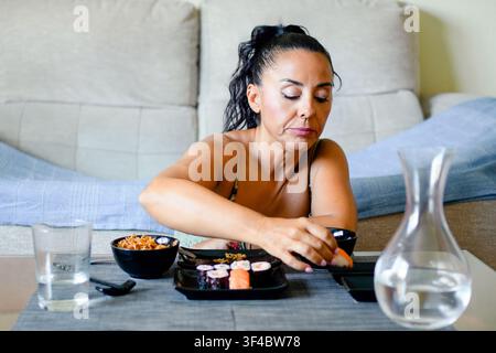 Donna rilassata mangia sushi in pace. Donna casual prende il sushi usando le bacchette a casa Foto Stock