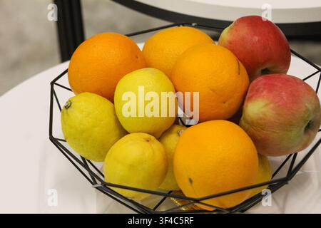 Arance, limoni e mele fresche in un moderno cesto di frutta in filo nero su un tavolo bianco. Concetto di cibo sano e decorazione domestica. Foto Stock