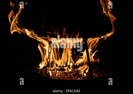 Fuoco di legna luminoso all'interno di una ciotola rotonda in metallo con fiamme ardenti nella notte buia. Vista ravvicinata dell'allestimento per cucinare in campeggio all'aperto con h Foto Stock