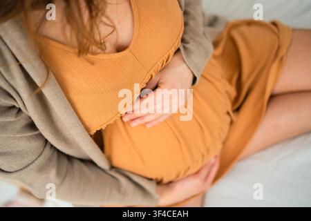 Una donna incinta siede su un divano bianco toccandosi la pancia in una stanza luminosa. La madre in attesa che accarezza il suo legame materno presenta un momento calmo e sereno della maternità naturale a casa. Foto Stock