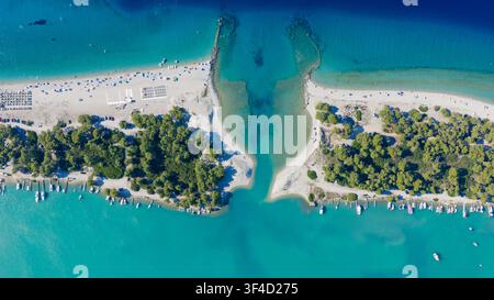 Vista dall'alto di una stretta laguna turchese che scorre tra rive ricoperte di alberi e una spiaggia sabbiosa allineata con ombrelloni e piccole barche. Foto Stock