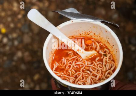 Una tazza di noodles istantanei piccanti con brodo rosso e cucchiaio di plastica bianco, zuppa saporita fumante e noodles intrecciati in un contenitore di carta monouso, sparsi casualmente Foto Stock