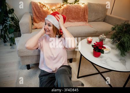 Donna con cappello di Babbo Natale seduta sul pavimento vicino al tavolino durante la stagione delle feste nel soggiorno Foto Stock