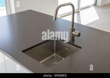 Una vista ravvicinata di un'isola cucina contemporanea con un piano di lavoro scuro, un lavello in acciaio inossidabile e un rubinetto minimalista Foto Stock