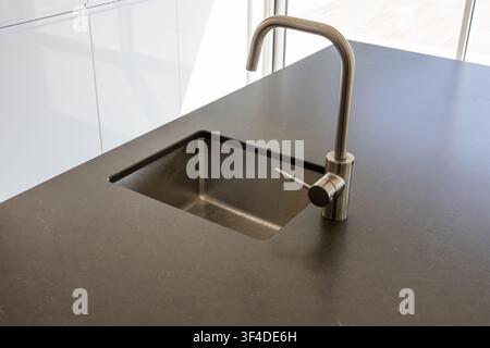 Una vista ravvicinata di un'isola cucina contemporanea con un piano di lavoro scuro, un lavello in acciaio inossidabile e un rubinetto minimalista Foto Stock