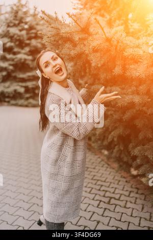 Donna che ride, autunnale elegante in cappotto a quadri in piedi accanto a pini in un parco illuminato dal sole che esprime gioia Foto Stock