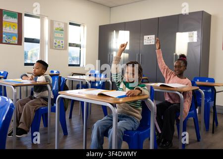 Studenti afroamericani seduti ai banchi nell'aula della scuola primaria con le mani alzate e i quaderni Foto Stock
