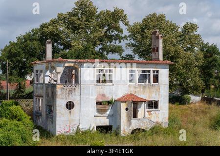 Antico edificio abbandonato a due piani con finestre rotte circondato da alberi verdi e vegetazione incolta Foto Stock