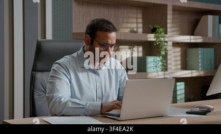 Uomo d'affari indiano arabo stanco e esausto che si tiene la testa, soffre di mal di testa, chiude il portatile dopo il lavoro, uomo d'affari in ufficio, lavoratore maschile che sente stanchezza e affaticamento agli occhi Foto Stock