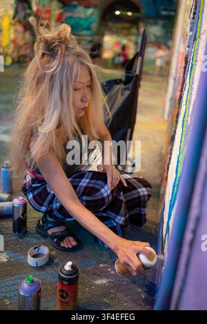 Artista graffiti maschile accovacciato che dipinge un murale colorato su un muro con bombole di vernice spray in un tunnel urbano, Londra. Foto Stock