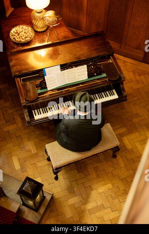 Vista ad angolo alto di un uomo con cappello fedora che suona un pianoforte a coda in una stanza pannellata in legno con spartiti e pavimentazione in parquet. Foto Stock