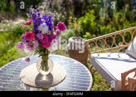 Prima piano di una composizione floreale messa su un tavolo di vetro in un giardino estivo. Zona lounge con mobili e mazzo di fiori freschi in una vaso Foto Stock