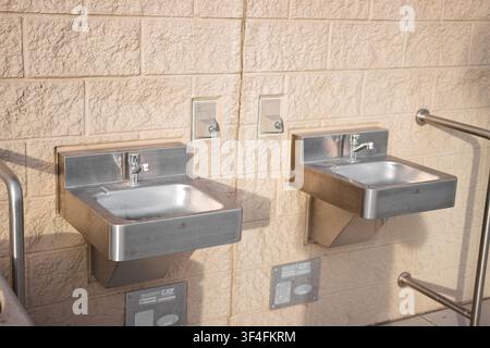Due lavandini esterni in acciaio inossidabile montati su un muro di cemento, progettati per il lavaggio pubblico delle mani con ringhiere di sicurezza e accesso pulito. Foto Stock