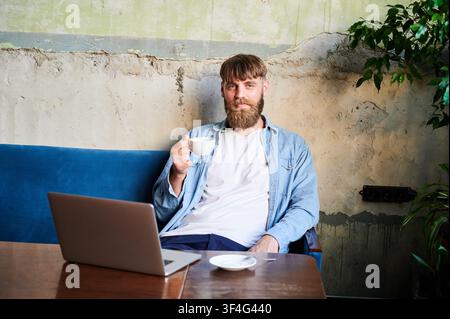 Ritratto di uomo barbuto con caffè seduto accanto a un laptop nell'interno di un caffè, sorridendo alla fotocamera in un momento di riflessione adatto per il lavoro da remoto, la consapevolezza, il pensiero creativo e i temi dello stile di vita Foto Stock