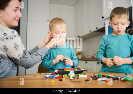 Madre che pulisce il naso di un bambino con un fazzoletto mentre due ragazzi giocano con blocchi di costruzione al tavolo. Un momento di genitorialità premurosa durante la vita familiare quotidiana a casa Foto Stock