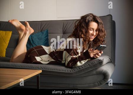 Donna sorridente sdraiata su un divano con smartphone, che mostra abitudini di comunicazione moderne, impegno sociale e stile di vita rilassato all'interno Foto Stock
