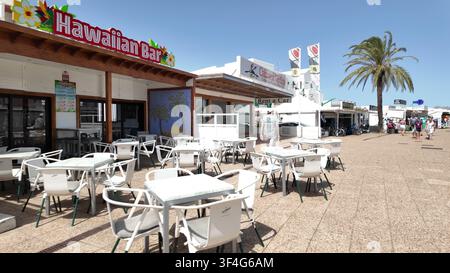 PUERTO DEL CARMEN, SPAGNA - 10 LUGLIO 2025: Tavoli e sedie bianchi all'aperto fuori da un bar hawaiano lungo la principale zona turistica di Puerto del Carmen, Lan Foto Stock
