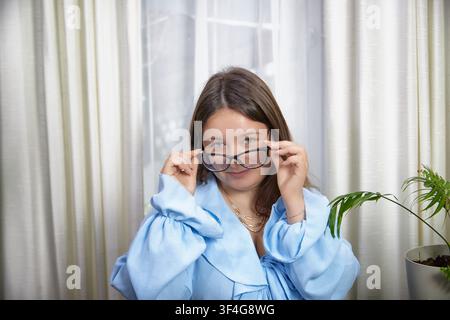 Donna adulta con occhiali in piedi vicino alla finestra in una casa luminosa, che mostra sicurezza e autoespressione in uno stile casual elegante Foto Stock