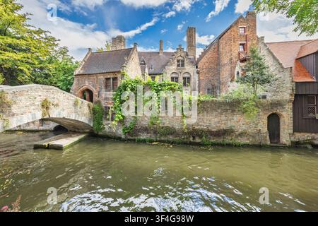 La città di Bruges in Belgio, Europa. Archi storici in pietra si inarcano su un canale accanto a vecchi edifici in mattoni adornati di lussureggiante edera verde sotto un blu vivace Foto Stock