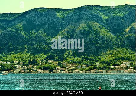 Un paesaggio orizzontale panoramico che mostra un villaggio mediterraneo costiero arroccato lungo un pendio montuoso lussureggiante, con case residenziali, tetti di tegole rosse, alberi fitti e piccole barche a vela bianche che scivolano sull'acqua calma in primo piano. Foto Stock