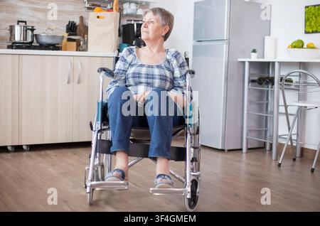 Donna anziana disabile pensierosa seduta su una sedia a rotelle che guarda attraverso una finestra della cucina, esprimendo solitudine, disabilità e tristezza del pensionamento. Foto Stock