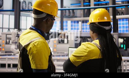 I membri del personale IT che sovrintendono alle attrezzature delle linee di produzione di pannelli solari. Colleghi che fanno brainstorming, mantenendo i sistemi di assemblaggio fotovoltaico in una fabbrica automatizzata dell'industria 4.0, telecamera A Foto Stock