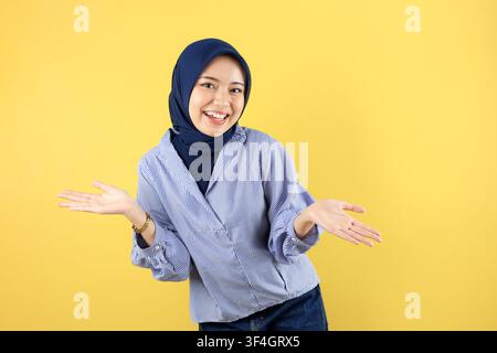Giovane donna musulmana indonesiana con hijab sembra felice con entrambe le mani sul fianco, isolata su sfondo giallo Foto Stock