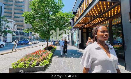 TORONTO, CANADA - 15 LUGLIO 2024: Una giovane donna cammina sul marciapiede passando davanti a negozi di lusso e luci di insegne sulla Bloor Street a Toronto, Canada. Foto Stock