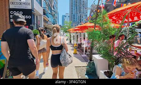 TORONTO, CANADA - 15 LUGLIO 2024: Persone camminano lungo una strada vivace fiancheggiata da caffè all'aperto e negozi nella zona di Yorkville a Toronto, Canada. Foto Stock