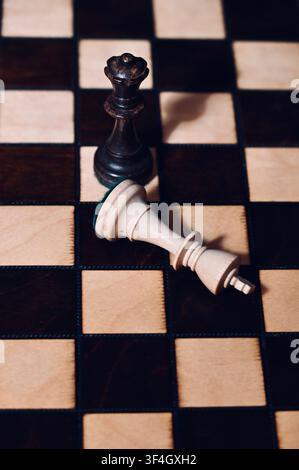 Una fotografia verticale dall'alto che mostra una regina nera in piedi sopra un re bianco caduto su una griglia di scacchi classica in legno. La scena simboleggia fortemente scacco matto, sconfitta, vittoria, cambio di potere, fine del gioco, successo tattico, leadership e strategia aziendale. Foto Stock
