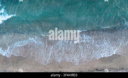 Vista aerea dall'alto con drone che cattura onde schiumose che rotolano sull'acqua azzurra verso una larga spiaggia di ciottoli a Nea Moudania, Grecia. Foto Stock