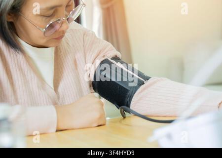 Donna asiatica di mezza età con occhiali che controlla la pressione a casa, monitor digitale. Routine quotidiana di cura della salute Foto Stock