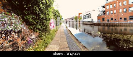 Sentiero del canale tranquillo fiancheggiato da vegetazione e mattoni coperti di graffiti, che riflette il paesaggio urbano. Camden, Londra, Regno Unito Foto Stock