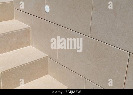 Una vista ravvicinata di gradini contemporanei in pietra beige che portano a un muro piastrellato con una piccola e discreta luce circolare Foto Stock