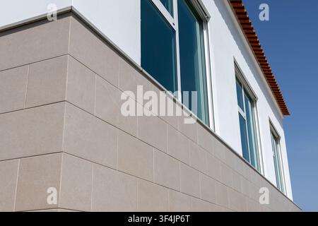 Una vista ravvicinata dell'angolo di un edificio mostra un rivestimento in pietra beige, finestre moderne e una parete superiore in stucco bianco contro un cielo blu limpido Foto Stock