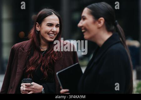 Due giovani amiche ridono davanti a un caffè mentre i colleghi chiacchierano con un tablet in città Foto Stock