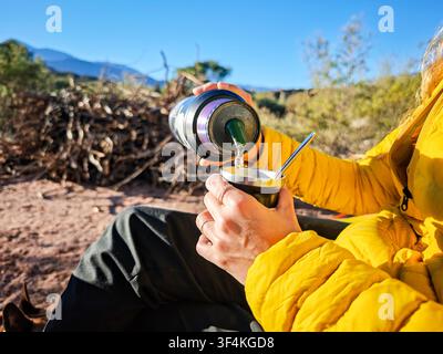 Donna che beve tè tradizionale alla yerba mate durante il campeggio in campagna in una giornata soleggiata Foto Stock