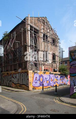 Magazzino storico in mattoni abbandonato, avvolto da impalcature e circondato da pannelli di compensato coperti di graffiti, Northern Quarter, Manchester, Regno Unito. Foto Stock