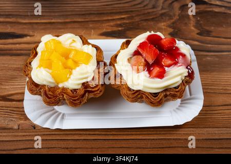 Due crostatine di sfoglia fragili ricoperte di panna, cubetti di mango fresco e fragole a fette su un piatto di carta bianco Foto Stock
