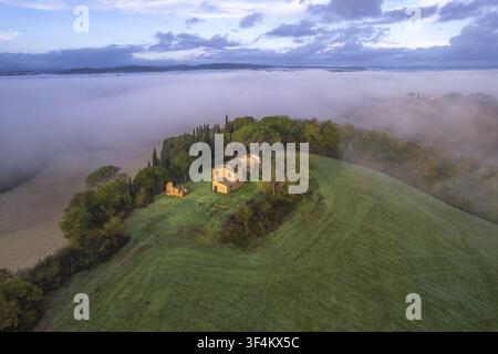 Vista aerea di una fattoria in pietra rustica circondata da cipressi e dolci colline verdi che emergono da una fitta nebbia mattutina nella campagna, Toscana Foto Stock